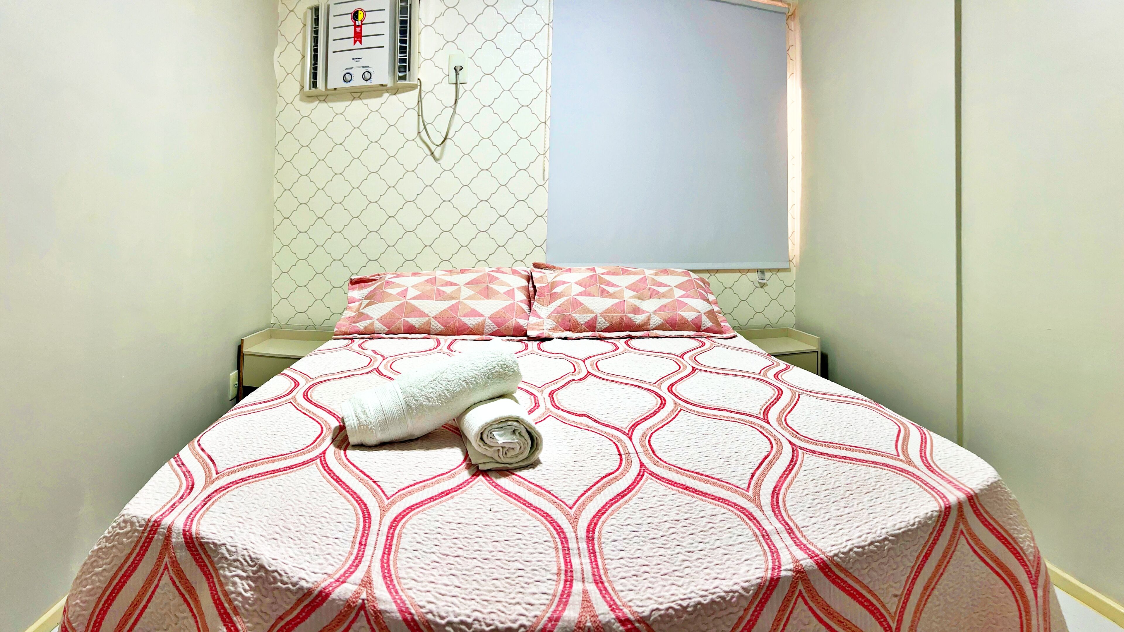 Una cama bien hecha con una colcha estampada, una toalla enrollada y un enchufe en la pared.