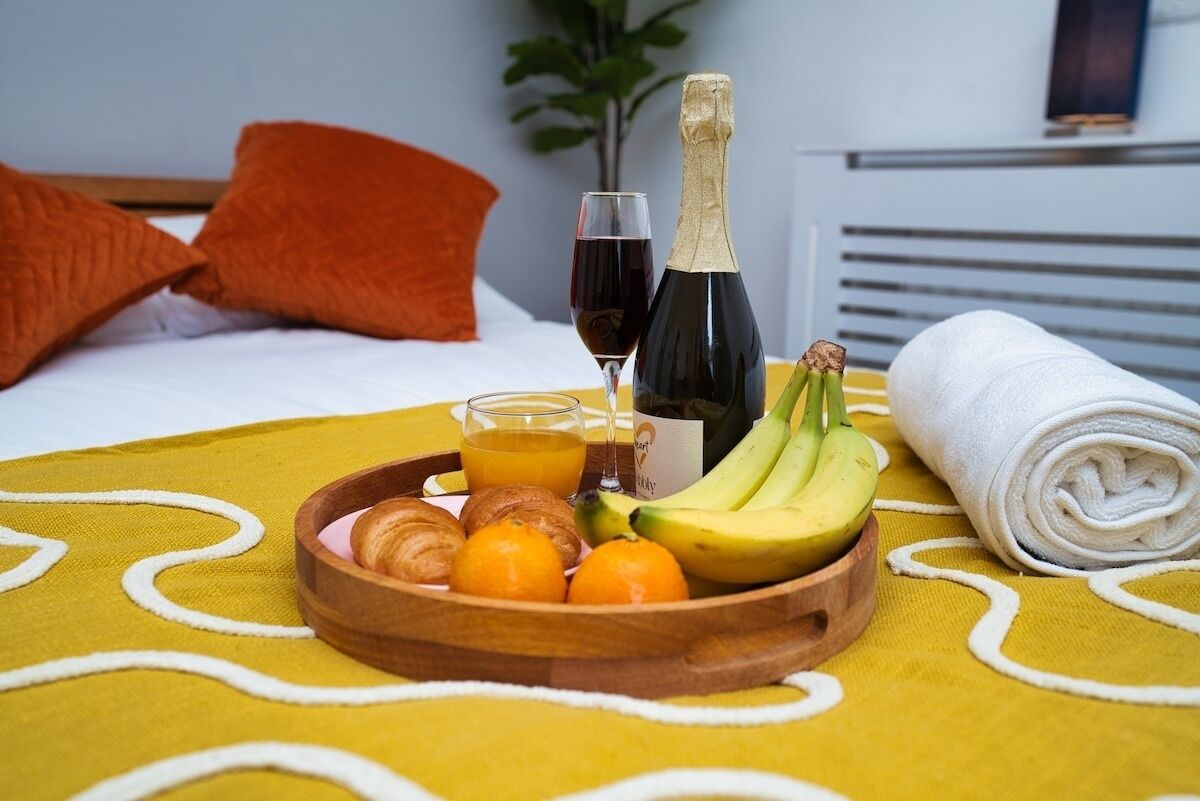 Ett frukostbricka med croissanter, bananer, apelsiner och juice, en flaska champagne och ett glas rött vin, placerat på en säng med ett gult överkast och orangefärgade kuddar.