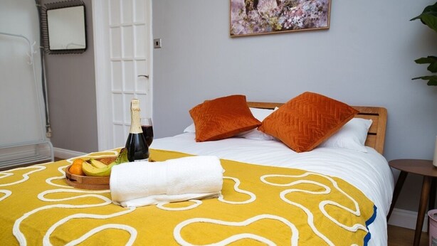 A neatly made bed with a yellow patterned bedspread, two orange pillows, a bottle of champagne, and a bowl of fruit. - Close to city centre home (Bradwell Common)