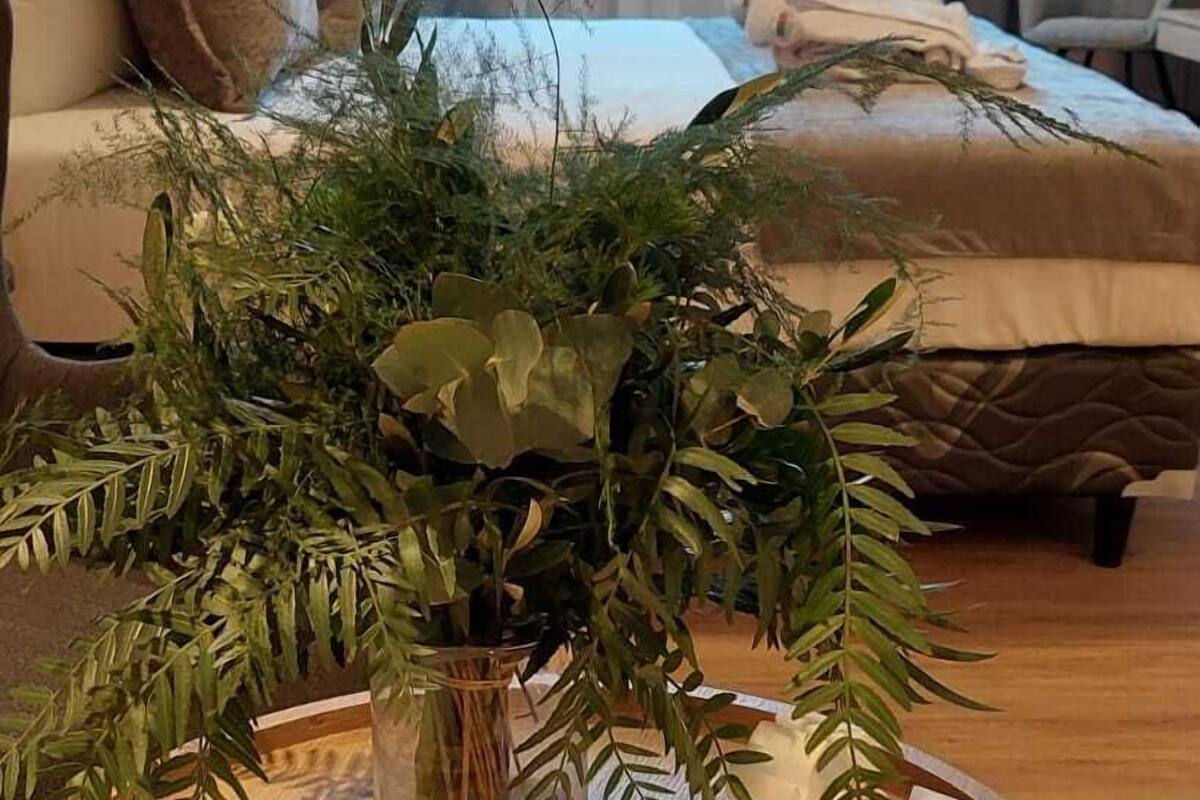 A round wooden table with a glass vase containing green foliage, a white decorative object, and a woven chair base.