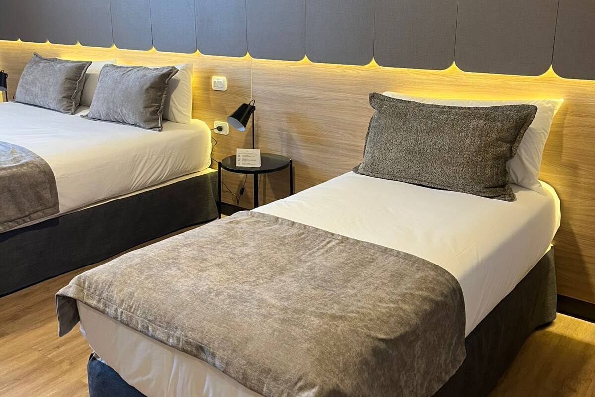 A hotel room with two beds, a nightstand, a lamp, and a wall-mounted light.