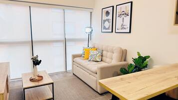 Una sala moderna con un sofá, una mesa de centro y una mesa de comedor. Hay dos cuadros enmarcados en la pared y un ventanal con persianas.