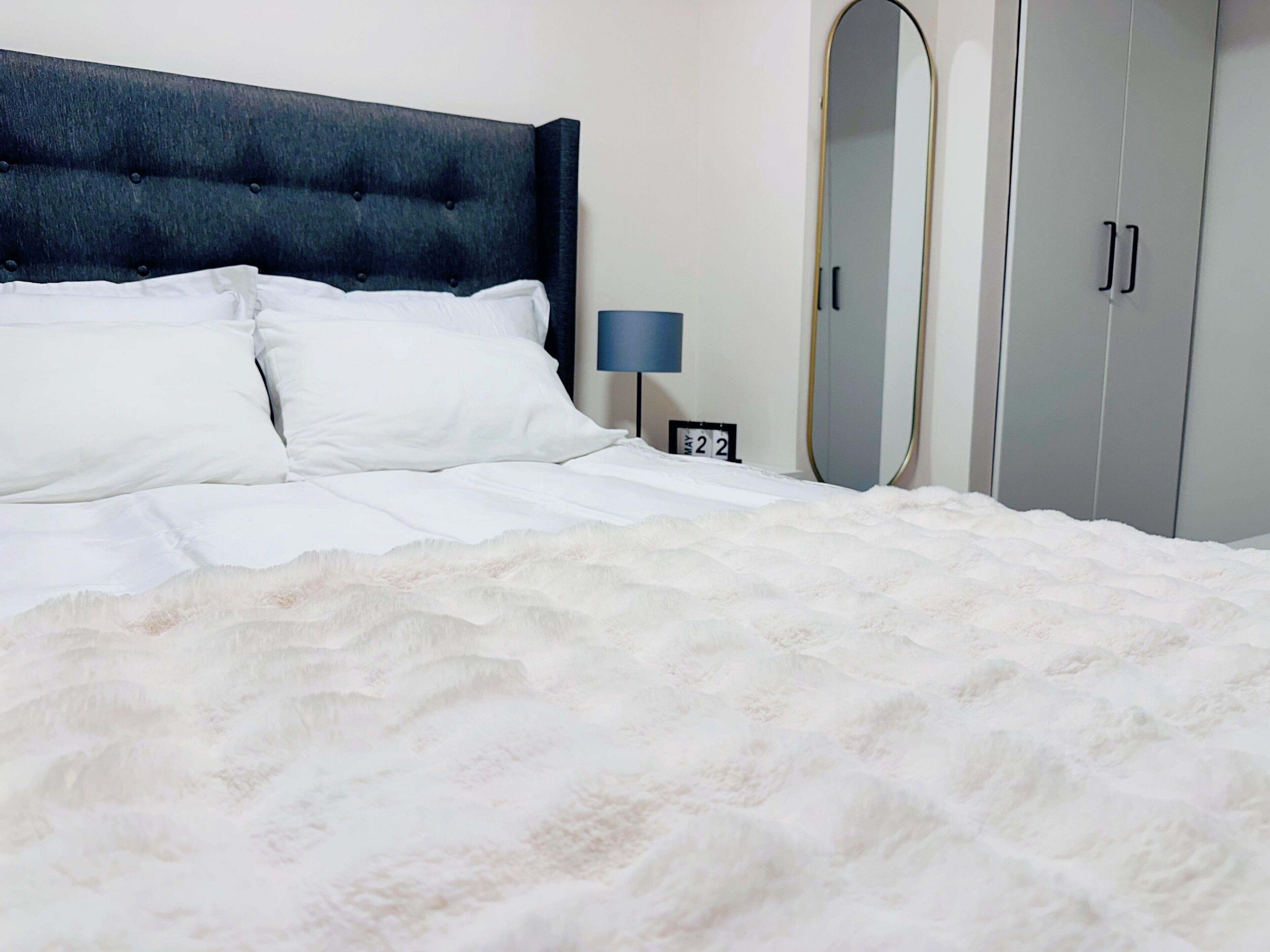 Una cama bien hecha con sábanas blancas y un edredón blanco mullido, un cabecero tapizado oscuro, una mesita de noche con una lámpara, un espejo y un armario con dos puertas.