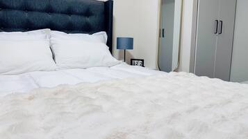 Una cama bien hecha con sábanas blancas y un edredón blanco mullido, un cabecero tapizado oscuro, una mesita de noche con una lámpara, un espejo y un armario con dos puertas.
