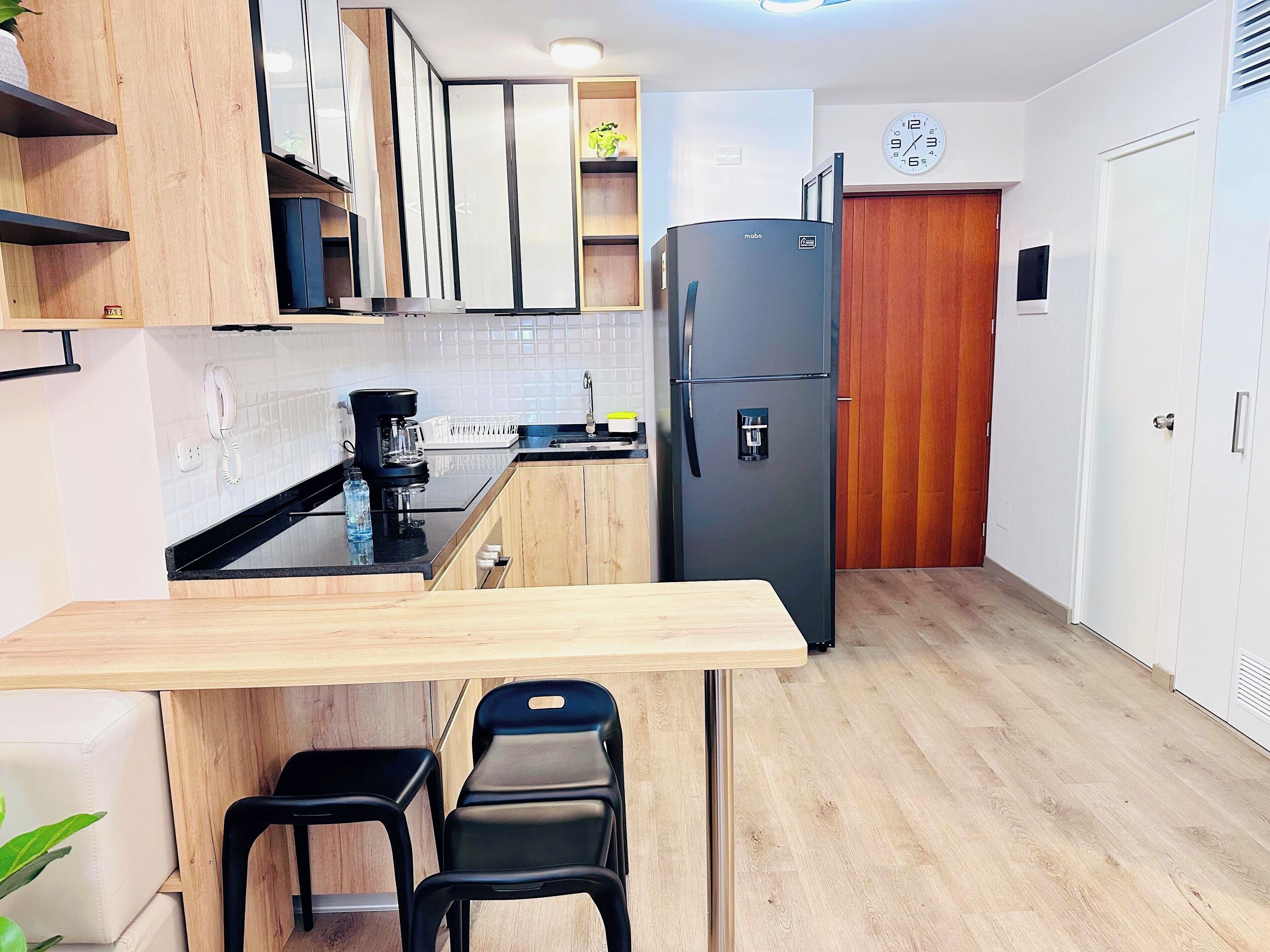 Una cocina moderna con armarios de madera, un refrigerador negro, una cafetera y un pequeño comedor con sillas.