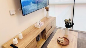 A modern living room with a mounted TV displaying a scenic view, a wooden console table with decorative items, and a cozy seating area with a woven basket and a vase of dried flowers.