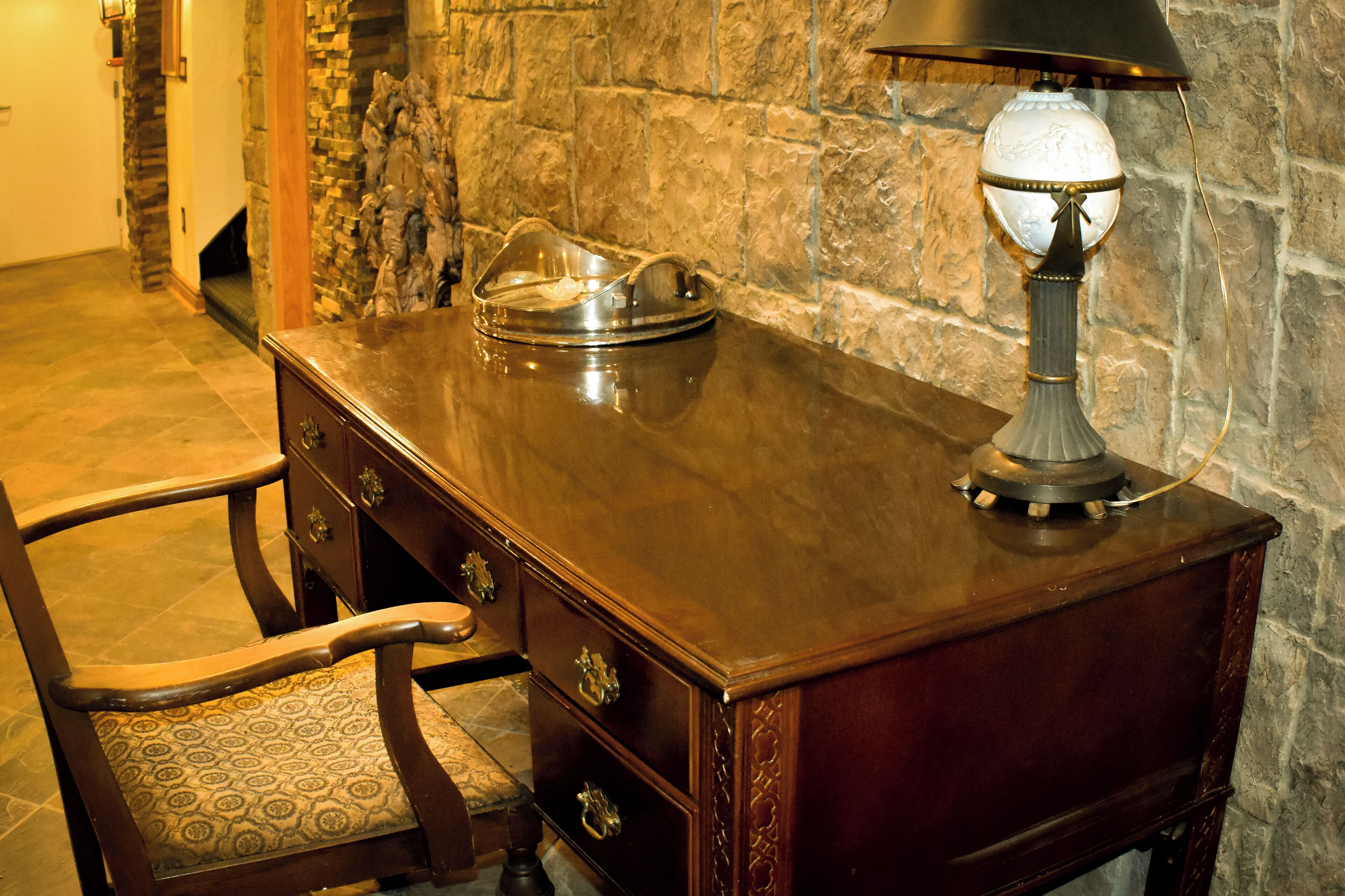 A wooden desk with drawers and a lamp, a chair with a patterned cushion, and a stone wall in the background.
