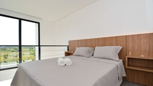 A modern bedroom with a large bed, a wooden headboard, and a view of greenery through large windows. - Loft - Haleiwa Novo Campeche (Florianopolis)