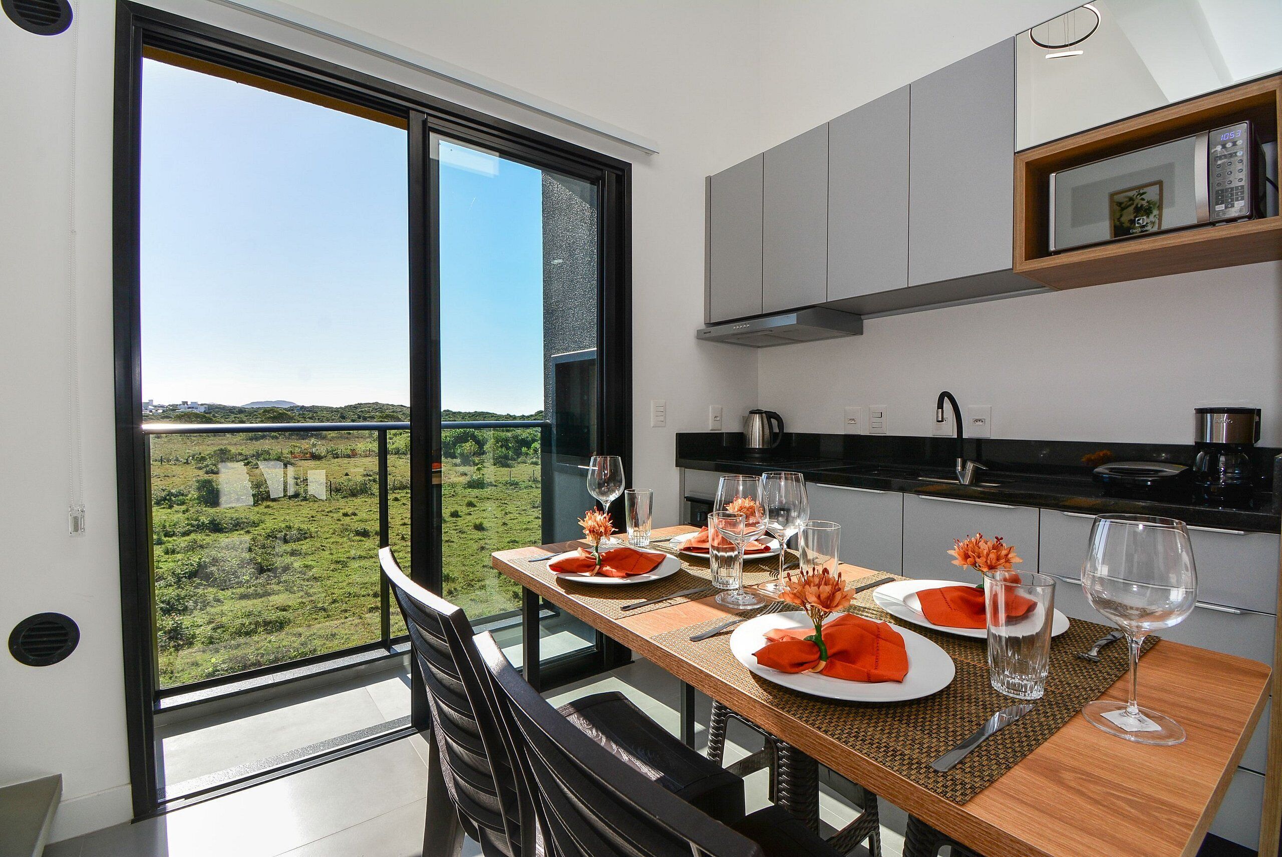 Un espace repas offrant une vue sur un paysage verdoyant grâce à de grandes fenêtres, une table en bois dressée pour deux personnes et des meubles de cuisine modernes.