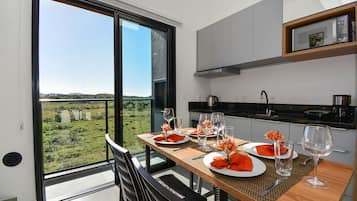 Un espace repas offrant une vue sur un paysage verdoyant grâce à de grandes fenêtres, une table en bois dressée pour deux personnes et des meubles de cuisine modernes.