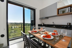 Comedor con vistas a un paisaje verde a través de amplios ventanales, una mesa de madera preparada para dos personas y modernos armarios de cocina.