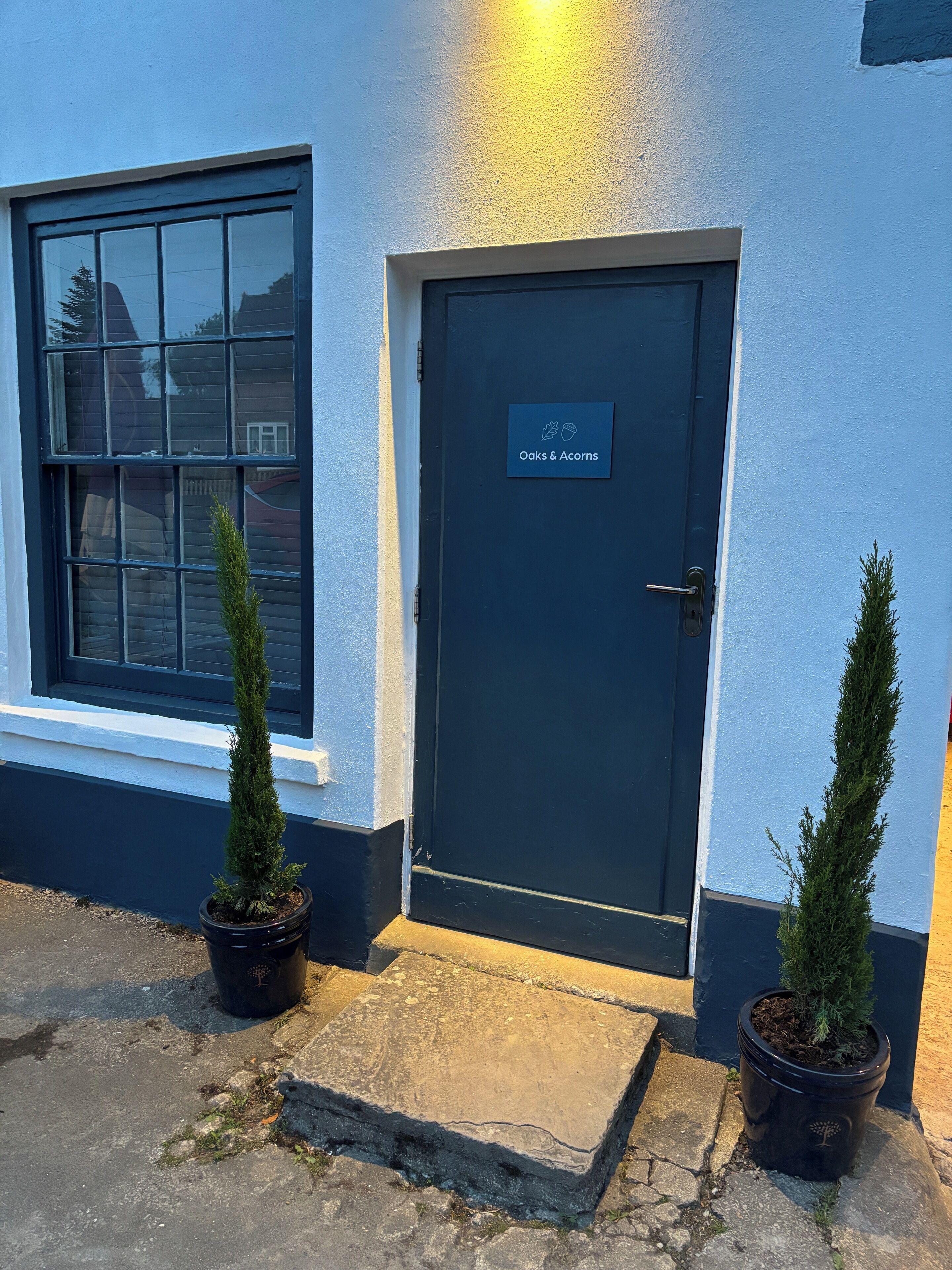 Una puerta azul oscuro con un cartel que dice "Oaks & Acorns" y dos arbustos perennes en macetas sobre la acera.