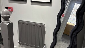 Een gang met een radiator, een kunstwerk in een lijst aan de muur, en een spiegel met een bijzondere golvende lijst.