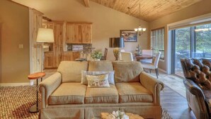 A cozy living room with a sofa, a coffee table, and a leather armchair. There is a dining area with a table and chairs, and a kitchen in the background.
