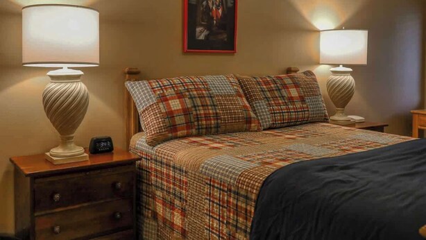 A bedroom with a plaid bedspread, two bedside lamps, a nightstand with a clock, and a framed picture on the wall.