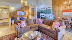 A modern living room with a brown sectional sofa, a glass coffee table, and a wooden staircase leading to a kitchen with teal cabinets.