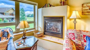 Un salon chaleureux avec une cheminée, une grande fenêtre et une vue sur une maison à l’extérieur.