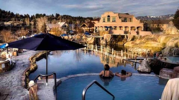 Un site de sources chaudes comprenant une piscine, une terrasse en bois et un grand bâtiment en arrière-plan.