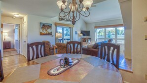 A dining area with a round table, chairs, a chandelier, and a view of the outdoors through large windows.