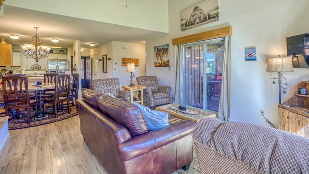 A spacious living area with a leather sofa, patterned armchairs, a dining table with chairs, a chandelier, and a large window with a view.