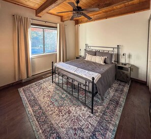 Un dormitorio con cama, ventilador de techo, ventana con cortinas y alfombra estampada.