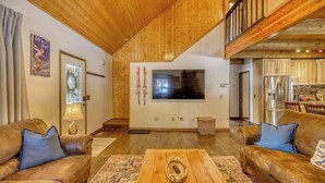 A spacious living room with wooden ceilings, a large TV, and a kitchen area visible through an open door. - Golf Course Hot Tub Cabin with Wildlife Views (Pagosa Springs)