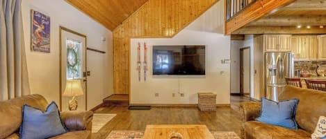 A spacious living room with wooden ceilings, a large TV, and a kitchen area visible through an open door.
