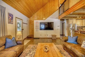 Amplia sala de estar con techos de madera, un televisor grande y una cocina visible a través de una puerta abierta.