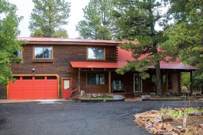 A two-story wooden cabin with a red garage door, a covered porch, and a stone foundation. - Golf Course Hot Tub Cabin with Wildlife Views (Pagosa Springs)
