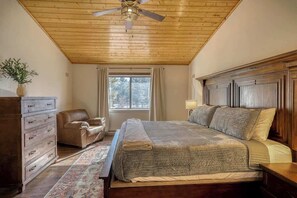 A bedroom with a wooden ceiling, a large bed with a headboard, a dresser, a chair, and a window with curtains.