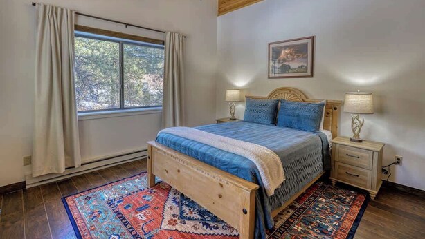 A bedroom with a wooden ceiling, a large window with curtains, a bed with a blue bedspread, two bedside tables with lamps, and a patterned rug. - Golf Course Hot Tub Cabin with Wildlife Views (Pagosa Springs)