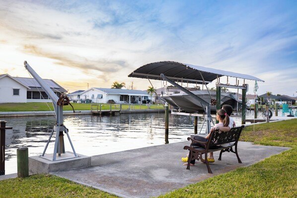 A bench under a covered area by a canal with houses in the background.