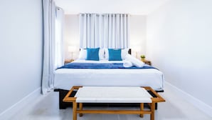 A neatly made bed with white and blue bedding, a wooden bench in front, and a small table with a lamp beside the bed.