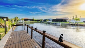 A wooden deck with a railing overlooking a calm body of water, boats docked in the background, and a clear sky.