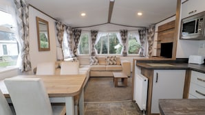A modern kitchen with a dining area, a built-in oven, and a seating area with a sofa and cushions. - Woodlands Gold (Ruthin)