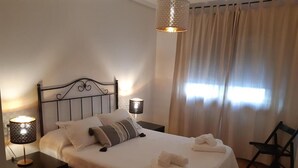 A neatly made bed with white linens and pillows, two bedside lamps, a chair, and a window with curtains.