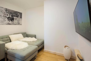 Habitación con sofá gris, un plaid blanco y mullido, un cuadro de ciervos, un televisor y una lámpara blanca.