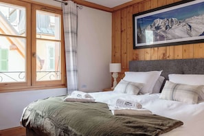 A hotel room with a bed, a window with a view of buildings, a painting of mountains, and wooden paneling.