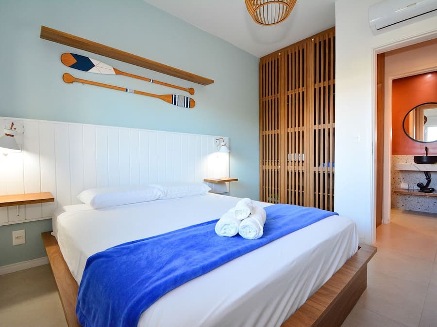 Una cama bien hecha con manta azul y almohadas blancas, cabecera de madera, y dos palas de madera colgadas en la pared.