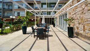 Ένα σκεπαστό, υπαίθριο αίθριο με τραπέζια και καρέκλες, ένας πέτρινος τοίχος και ένα πολυώροφο κτίριο στο βάθος.