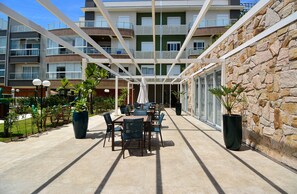A covered outdoor patio with tables and chairs, a stone wall, and a multi-story building in the background. - SunClub-300 metros da Praia do Campeche (Florianopolis)