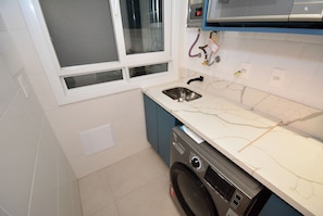 A compact kitchen with a built-in washing machine, a window with blinds, and a modern countertop. - SunClub-300 metros da Praia do Campeche (Florianopolis)
