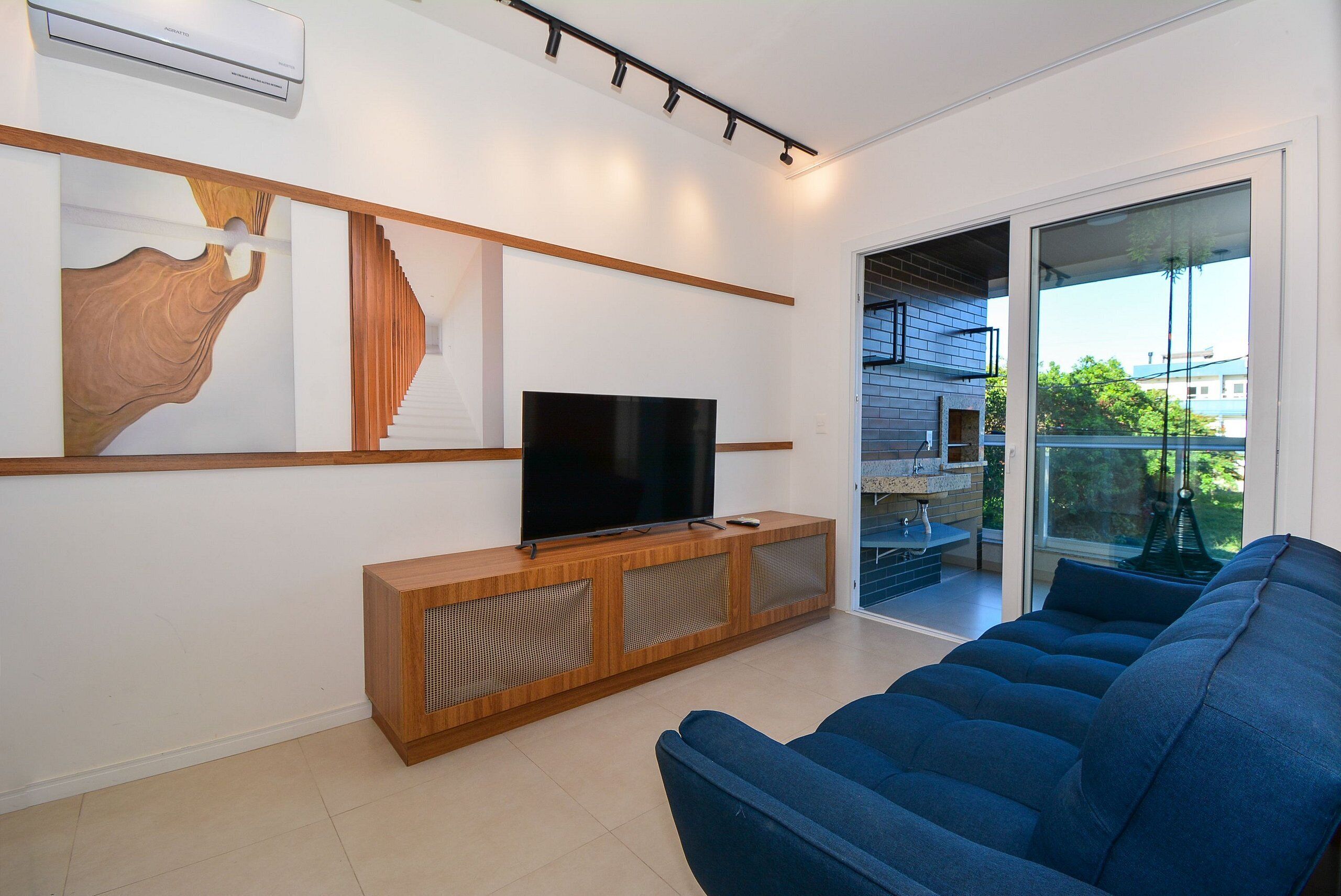 Een moderne woonkamer met een flatscreen televisie op een houten console, een blauwe bank en een groot raam met uitzicht.