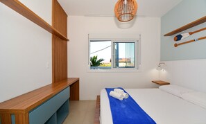 Une chambre à coucher comprenant un lit, un banc en bois, une fenêtre et une étagère fixée au mur, sur laquelle reposent deux battes de bois.
