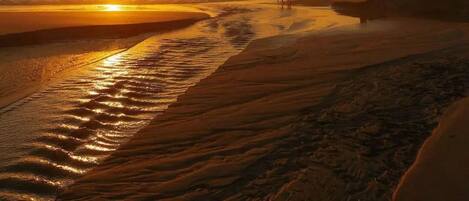 A beach scene at sunset with a person and a dog walking along the shore.