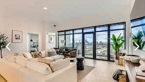 A modern living room with a large white sofa, a coffee table, and a view of the city through floor-to-ceiling windows. - Luxury penthouse in enchanting Denver with bar, gym, pool, sauna & more (Denver)