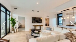 A modern living room with a large sofa, a coffee table, and a TV mounted on the wall. There is a plant and a window with blinds. - Luxury penthouse in enchanting Denver with bar, gym, pool, sauna & more (Denver)