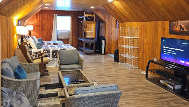 A spacious loft-style living area with high ceilings, wooden beams, and a cozy seating arrangement including a sofa and chairs. A television is mounted on the wall, and there is a small table with a lamp. - Cabin Style Private Studio  (Giroux)