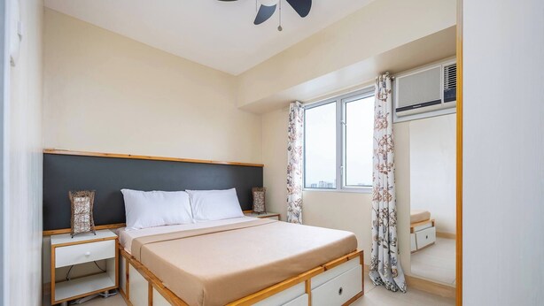 A bedroom with a bed, nightstands, a dresser, a window with curtains, and a ceiling fan. - 2-bedroom Retreat in Avida Riala Prime Location (Cebu City)