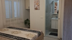A bedroom with a bed, a window with blinds, a potted plant, and a bathroom visible through an open door.